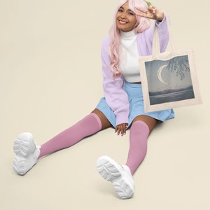 May include: A person with pink hair is sitting on the floor with their legs crossed. They are wearing a light purple cardigan, a white turtleneck, a light blue skirt, and pink knee-high socks. They are holding a white tote bag with a crescent moon and a tree design. The bag has a light blue, gray, and white color scheme.