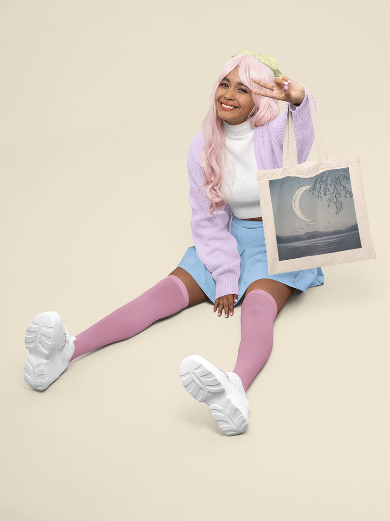 May include: A person with pink hair is sitting on the floor with their legs crossed. They are wearing a light purple cardigan, a white turtleneck, a light blue skirt, and pink knee-high socks. They are holding a white tote bag with a crescent moon and a tree design. The bag has a light blue, gray, and white color scheme.
