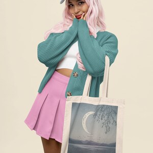 May include: A woman wearing a teal cardigan, pink skirt, and white cap is holding a tote bag with a crescent moon and tree illustration.