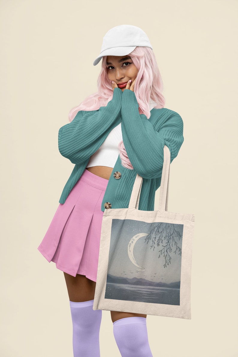 May include: A woman wearing a teal cardigan, pink skirt, and white cap is holding a tote bag with a crescent moon and tree illustration.