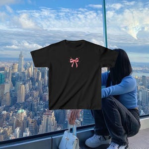 May include: Black t-shirt with a pink bow graphic on the front. The shirt is being worn by a person standing in front of a window with a city view.