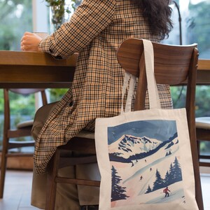 以下が含まれることがあります： スキーヤーが描かれた雪山風景のカラフルなイラストが施された白いトートバッグ。バッグには長いストラップが付いており、木製の椅子の背もたれにかけられています。