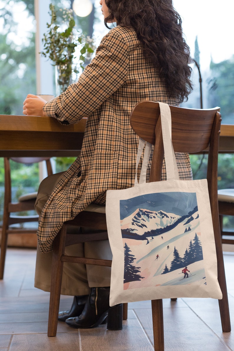 以下が含まれることがあります： スキーヤーが描かれた雪山風景のカラフルなイラストが施された白いトートバッグ。バッグには長いストラップが付いており、木製の椅子の背もたれにかけられています。