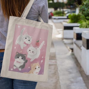 May include: A canvas tote bag featuring a pink background with illustrations of four adorable cartoon kittens. The kittens are in various poses, with some wearing bow ties and surrounded by stars and a small apple. The bag is carried over a shoulder.