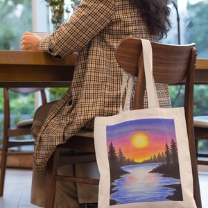 May include: A white tote bag with a colorful sunset scene painted on it. The scene features a lake, trees, and a bright orange sun.