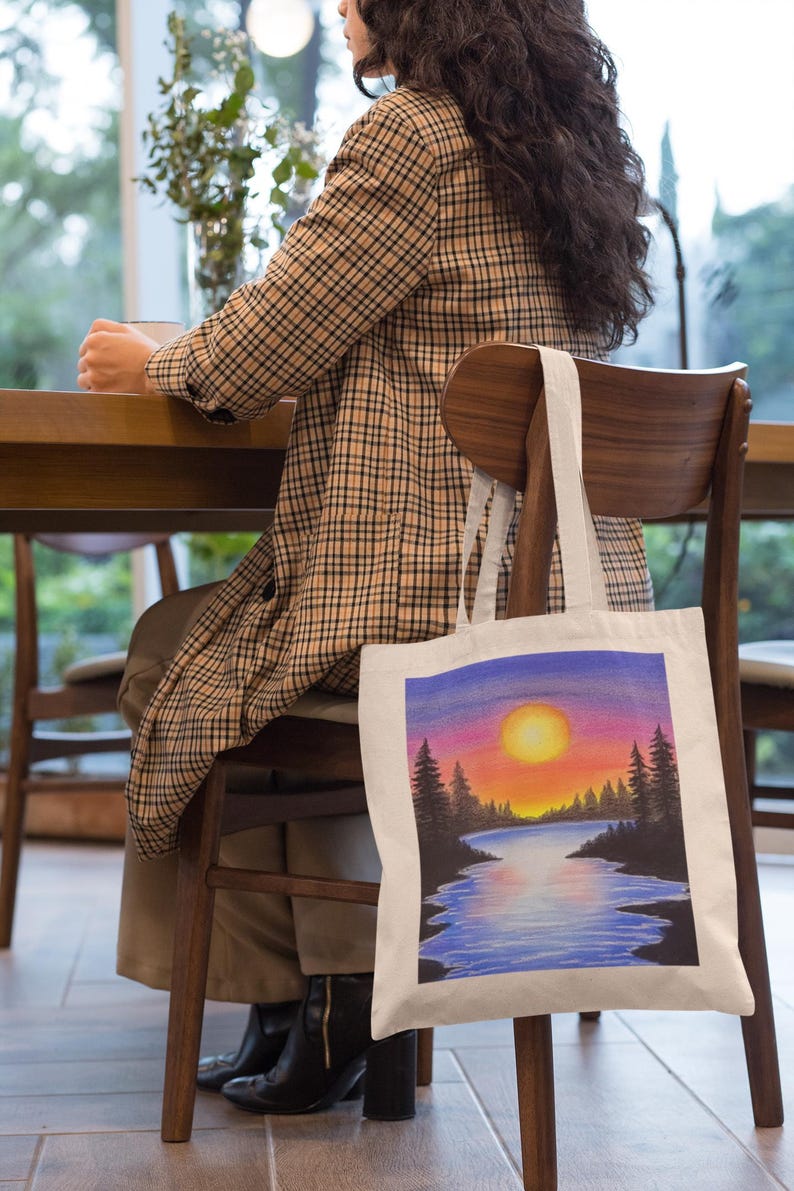 May include: A white tote bag with a colorful sunset scene painted on it. The scene features a lake, trees, and a bright orange sun.