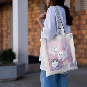 May include: A canvas tote bag featuring a pink background with illustrations of five cartoon cats. The cats are in various poses, with a small red apple graphic. The bag has a white strap and is being carried.