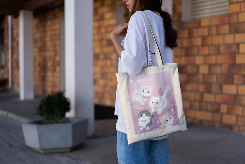 May include: A canvas tote bag featuring a pink background with illustrations of five cartoon cats. The cats are in various poses, with a small red apple graphic. The bag has a white strap and is being carried.