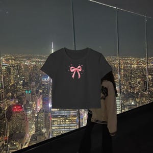 May include: Black cropped t-shirt with a pink bow and hearts design. The shirt is photographed against a city skyline at night.
