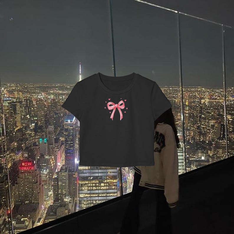 May include: Black cropped t-shirt with a pink bow and hearts design. The shirt is photographed against a city skyline at night.