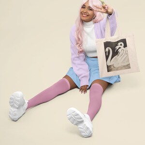 May include: Beige tote bag featuring a black and white illustration of swans. The bag is held by a person wearing a light purple cardigan, a white turtleneck, and a blue skirt. The person is also wearing pink knee-high socks and white sneakers.