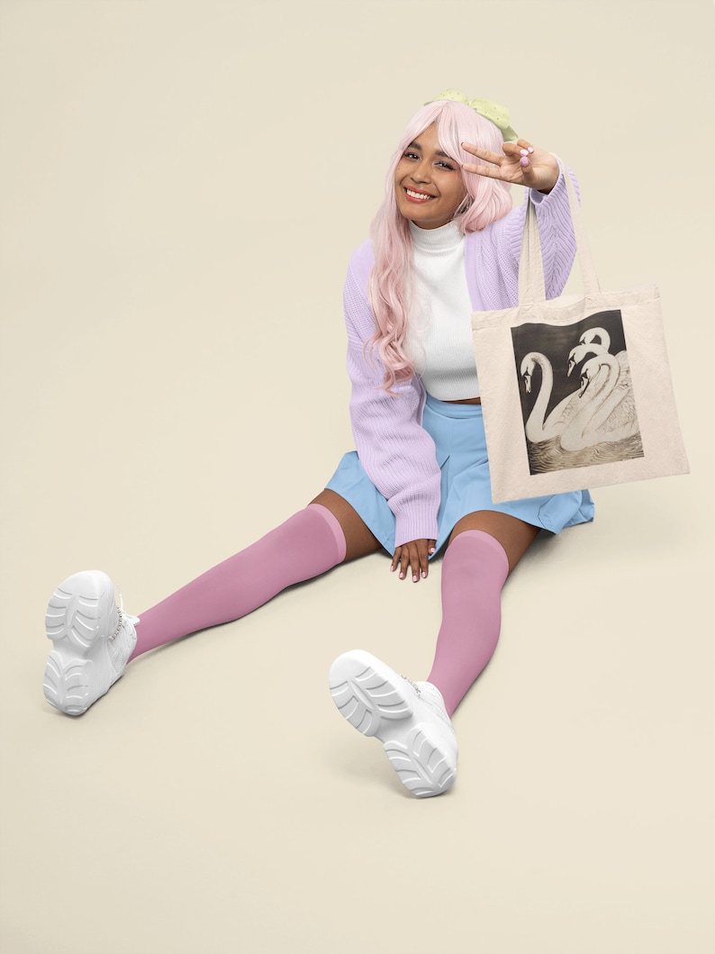 May include: Beige tote bag featuring a black and white illustration of swans. The bag is held by a person wearing a light purple cardigan, a white turtleneck, and a blue skirt. The person is also wearing pink knee-high socks and white sneakers.