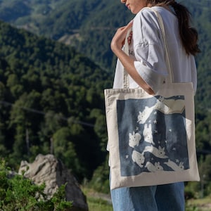 May include: A white canvas tote bag with a blue design of white rabbits jumping in the air with stars. The rabbits are outlined in black and have black eyes. The bag has two white straps.