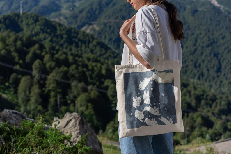 May include: A white canvas tote bag with a blue design of white rabbits jumping in the air with stars. The rabbits are outlined in black and have black eyes. The bag has two white straps.