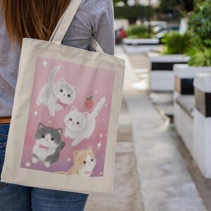 May include: A canvas tote bag featuring a pink background with illustrations of four adorable cartoon kittens. The kittens are in various poses, with some wearing bow ties and surrounded by stars and a small apple. The bag is carried over a shoulder.