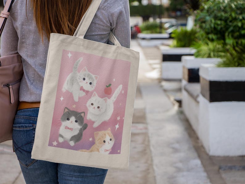 May include: A canvas tote bag featuring a pink background with illustrations of four adorable cartoon kittens. The kittens are in various poses, with some wearing bow ties and surrounded by stars and a small apple. The bag is carried over a shoulder.