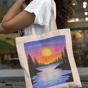 May include: A white canvas tote bag with a colorful sunset scene painted on it. The scene features a lake with a large orange sun setting behind a line of trees. The sky is a gradient of purple, pink, and orange.