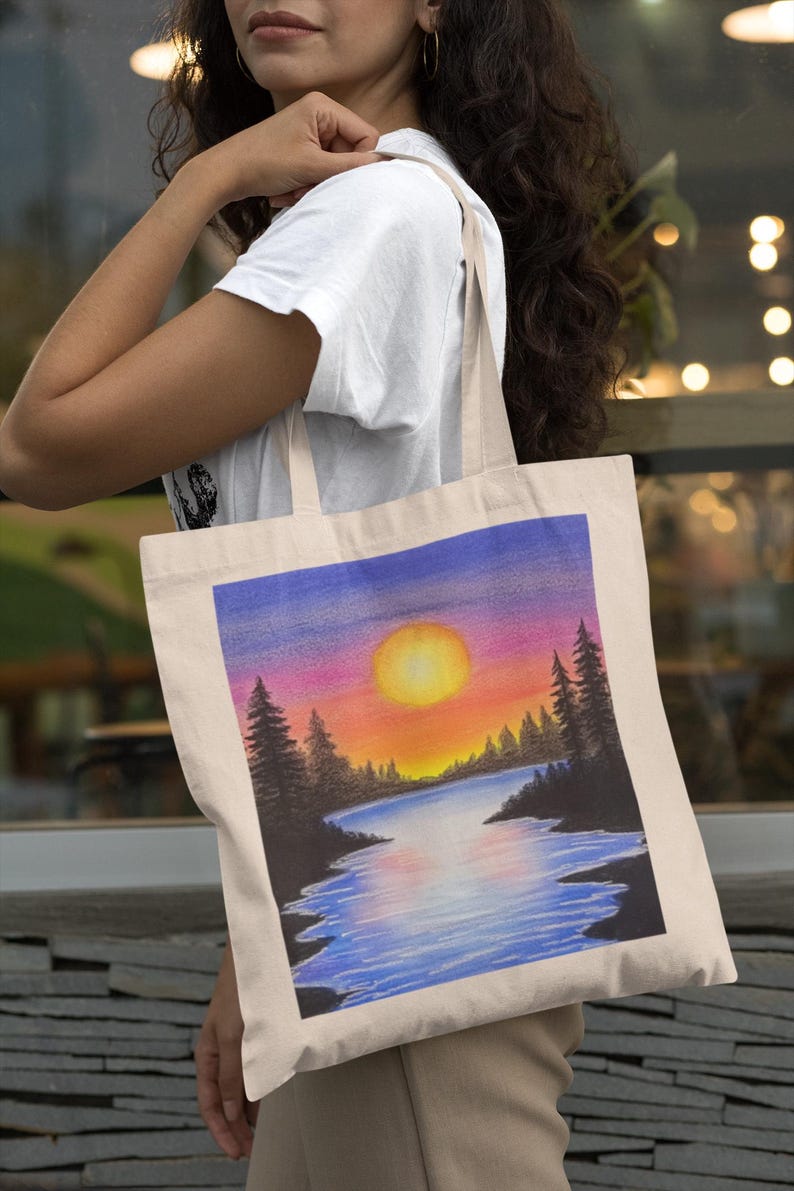 May include: A white canvas tote bag with a colorful sunset scene painted on it. The scene features a lake with a large orange sun setting behind a line of trees. The sky is a gradient of purple, pink, and orange.