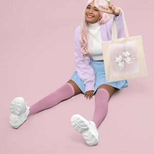 May include: A young person with pink hair wearing a light purple cardigan, a white turtleneck, a light blue skirt, and pink knee-high socks. They are sitting on a pink background and holding a white tote bag with a pink and white illustration of two cats. The bag has a light brown handle.
