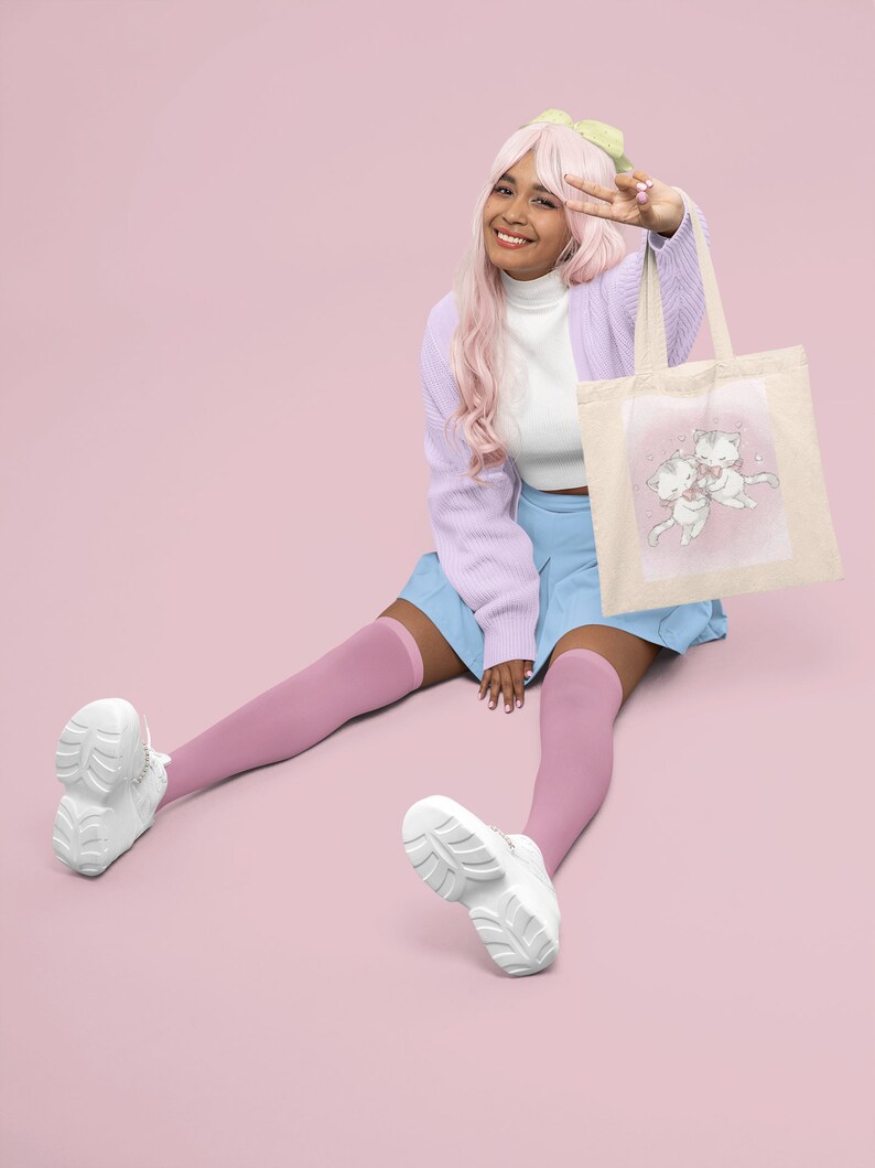 May include: A young person with pink hair wearing a light purple cardigan, a white turtleneck, a light blue skirt, and pink knee-high socks. They are sitting on a pink background and holding a white tote bag with a pink and white illustration of two cats. The bag has a light brown handle.