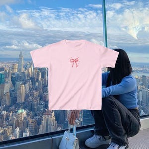 May include: A pink t-shirt with a pink bow and small hearts on the front. The shirt is being worn by a person standing in front of a window with a city view.