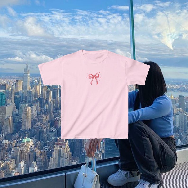 May include: A pink t-shirt with a pink bow and small hearts on the front. The shirt is being worn by a person standing in front of a window with a city view.
