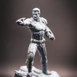 May include: A grey painted figurine of a superhero standing on a rocky base. The superhero is wearing a suit with a belt and is holding a weapon in their right hand.