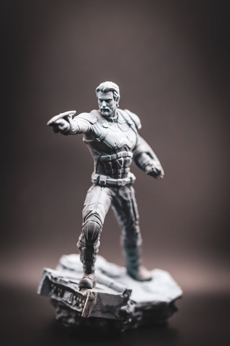 May include: A grey painted figurine of a superhero standing on a rocky base. The superhero is wearing a suit with a belt and is holding a weapon in their right hand.