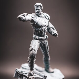 May include: A grey painted figurine of a superhero standing on a grey, rocky base. The superhero is wearing a detailed costume with a belt and is holding a weapon in their right hand.