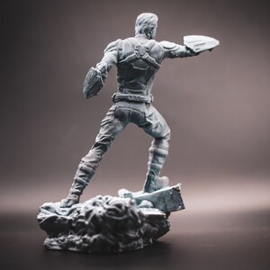May include: A grey plastic figurine of a superhero standing on a rocky base. The superhero is wearing a blue and red costume and is holding a shield in one hand.