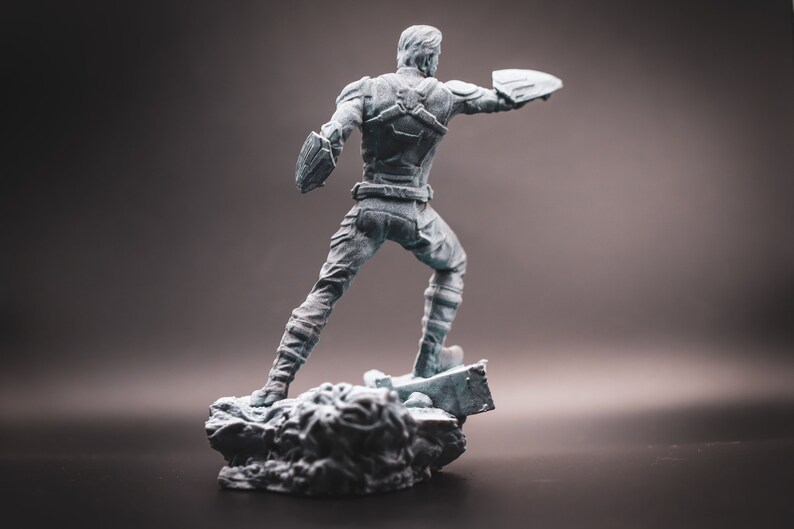 May include: A grey plastic figurine of a superhero standing on a rocky base. The superhero is wearing a blue and red costume and is holding a shield in one hand.