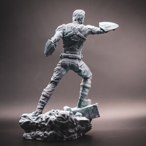 May include: A grey plastic figurine of a superhero standing on a rocky base. The superhero is wearing a suit with a cape and holding a shield in one hand.