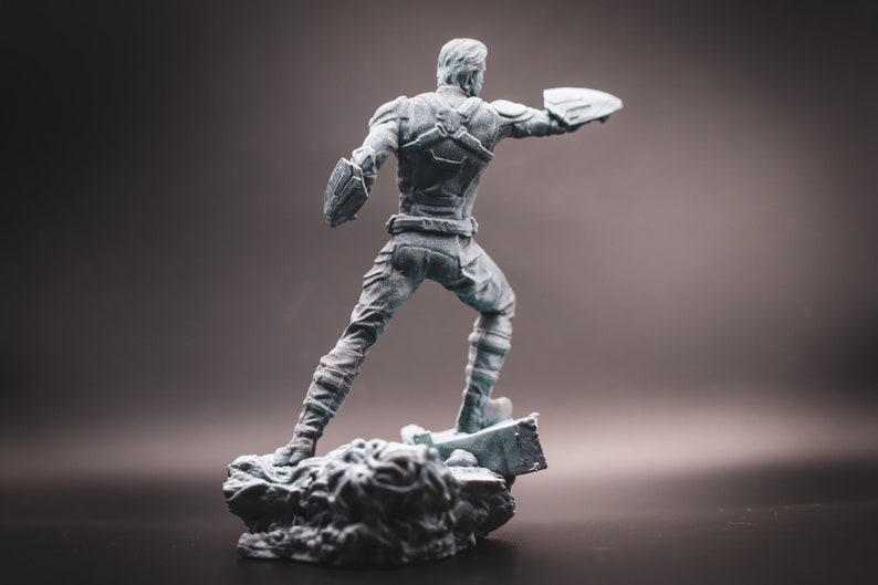 May include: A grey plastic figurine of a superhero standing on a rocky base. The superhero is wearing a suit with a cape and holding a shield in one hand.