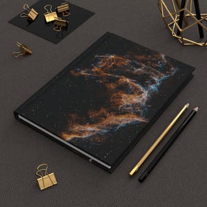 Op de afbeelding: Een zwart notitieboek met een nevelontwerp in oranje, blauw en wit. Gouden en zwarte potloden en gouden paperclips zijn ook aanwezig. Het thema is briefpapier en kantoorbenodigdheden.