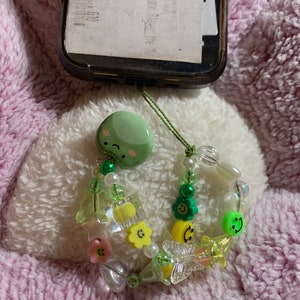 以下が含まれることがあります： 緑色のスマイリーフェイスチャームと緑、ピンク、黄色の配色が施されたビーズの携帯電話チャーム。チャームには、さまざまなビーズ、花、星、蝶が含まれています。携帯電話ケースは黒です。