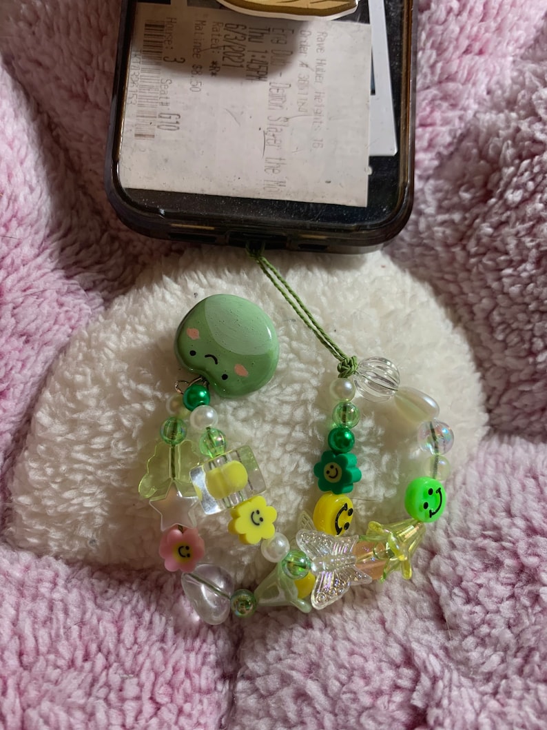 以下が含まれることがあります： 緑色のスマイリーフェイスチャームと緑、ピンク、黄色の配色が施されたビーズの携帯電話チャーム。チャームには、さまざまなビーズ、花、星、蝶が含まれています。携帯電話ケースは黒です。
