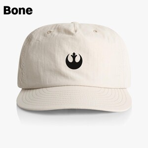 May include: A beige baseball cap with a black Rebel Alliance symbol on the front. The cap has a curved brim and a structured crown.
