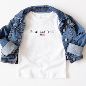 May include: A white t-shirt with the words "feral and free" and a small American flag graphic. The shirt is paired with a blue denim jacket. The jacket is folded over the shirt.