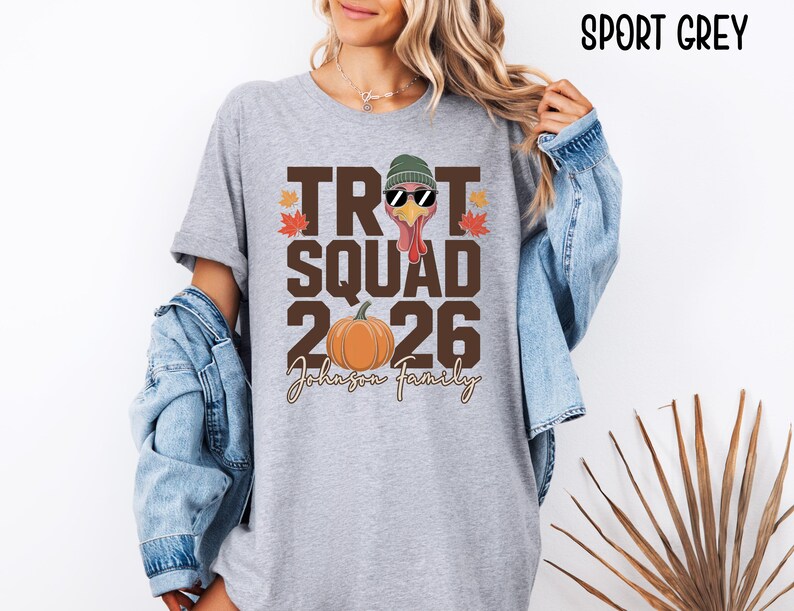 May include: A sport grey t-shirt featuring a graphic with the text TROT SQUAD 2026 and Johnson Family. The design includes a turkey wearing a green beanie and sunglasses, an orange pumpkin, and red and orange maple leaves on a grey background.