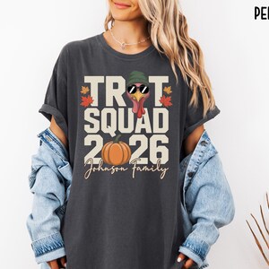 May include: A charcoal gray t-shirt featuring a graphic print with the text TROT SQUAD 2026 and Johnson Family. The design includes a turkey wearing a green beanie and sunglasses, an orange pumpkin, and colorful autumn leaves on the sides.