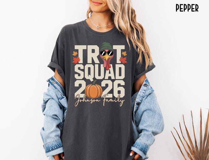 May include: A charcoal gray t-shirt featuring a graphic print with the text TROT SQUAD 2026 and Johnson Family. The design includes a turkey wearing a green beanie and sunglasses, an orange pumpkin, and colorful autumn leaves on the sides.