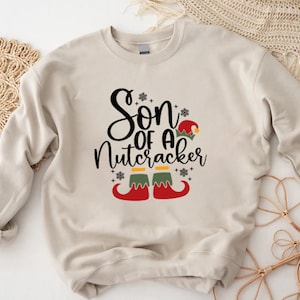 May include: A beige sweatshirt with the text "Son of a Nutcracker" in black lettering with a green and red elf shoe graphic.