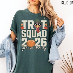 May include: A blue spruce colored t-shirt featuring a graphic of a turkey wearing a green beanie and sunglasses, with the text TROT SQUAD 2026 and Johnson Family. The design includes a pumpkin and autumn leaves in orange, red, and yellow colors.