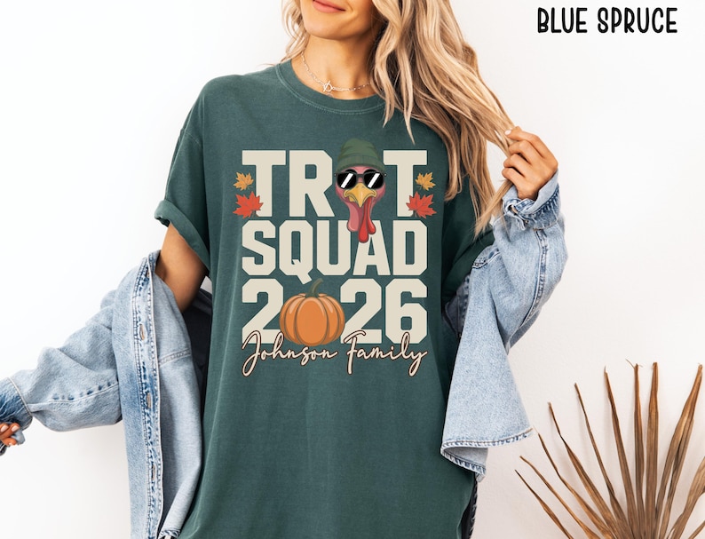 May include: A blue spruce colored t-shirt featuring a graphic of a turkey wearing a green beanie and sunglasses, with the text TROT SQUAD 2026 and Johnson Family. The design includes a pumpkin and autumn leaves in orange, red, and yellow colors.