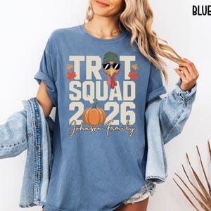 May include: A blue jean colored t-shirt featuring a graphic of a turkey wearing a green beanie and sunglasses. The text reads TROT SQUAD 2026 and Johnson Family, accompanied by an orange pumpkin and small red and yellow autumn leaf illustrations.