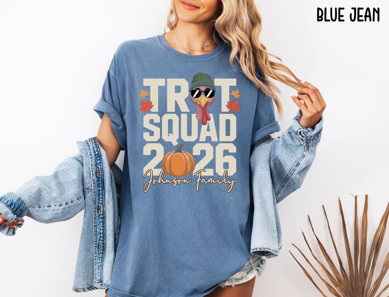 May include: A blue jean colored t-shirt featuring a graphic of a turkey wearing a green beanie and sunglasses. The text reads TROT SQUAD 2026 and Johnson Family, accompanied by an orange pumpkin and small red and yellow autumn leaf illustrations.