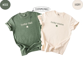 May include: Two t-shirts, one moss green and one ivory, are displayed. The moss green shirt says "lucky dad" with a shamrock and "Est. 2026" on the sleeve. The ivory shirt says "lucky mama" with a shamrock and "Est. 2026" on the sleeve. The word "customizable" is above the green shirt.