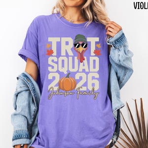May include: A violet t-shirt featuring a graphic of a turkey wearing a green beanie and black sunglasses. The text reads TROT SQUAD 2026 and Johnson Family, accompanied by an orange pumpkin and small red and yellow autumn leaves.