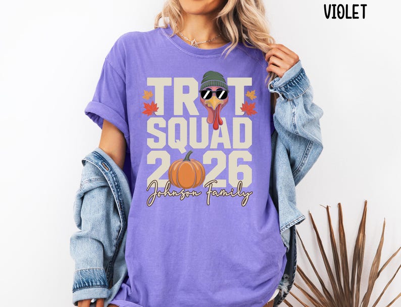 May include: A violet t-shirt featuring a graphic of a turkey wearing a green beanie and black sunglasses. The text reads TROT SQUAD 2026 and Johnson Family, accompanied by an orange pumpkin and small red and yellow autumn leaves.