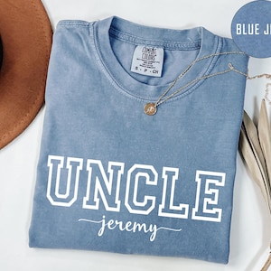 May include: A blue jean-colored t-shirt with the word "UNCLE" in white block letters and the name "jeremy" in script. A gold necklace with a pendant is draped across the shirt. The shirt is folded on a white surface.